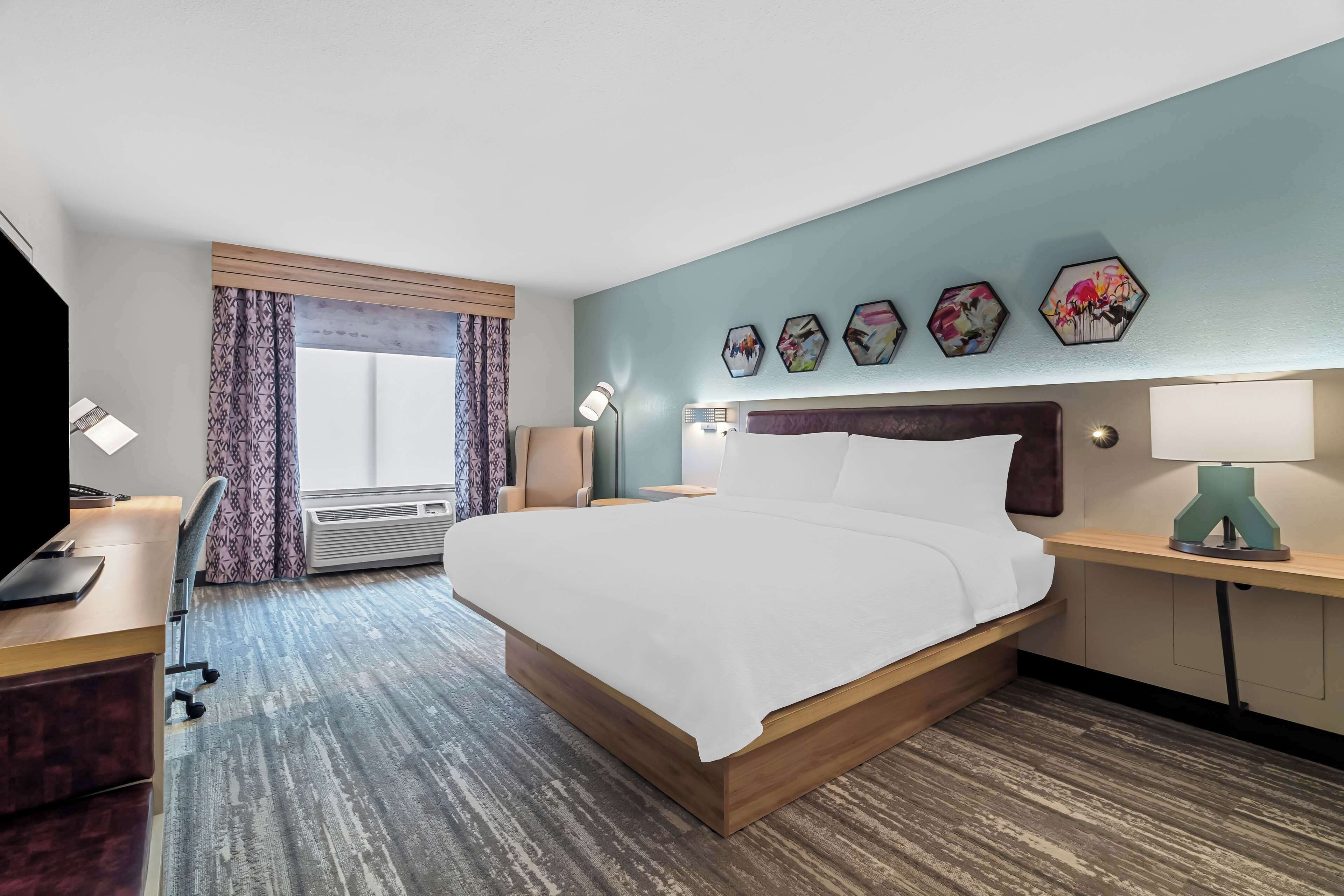 A hotel room with a large bed, a desk with a computer, a chair, a desk lamp, and a window with curtains.