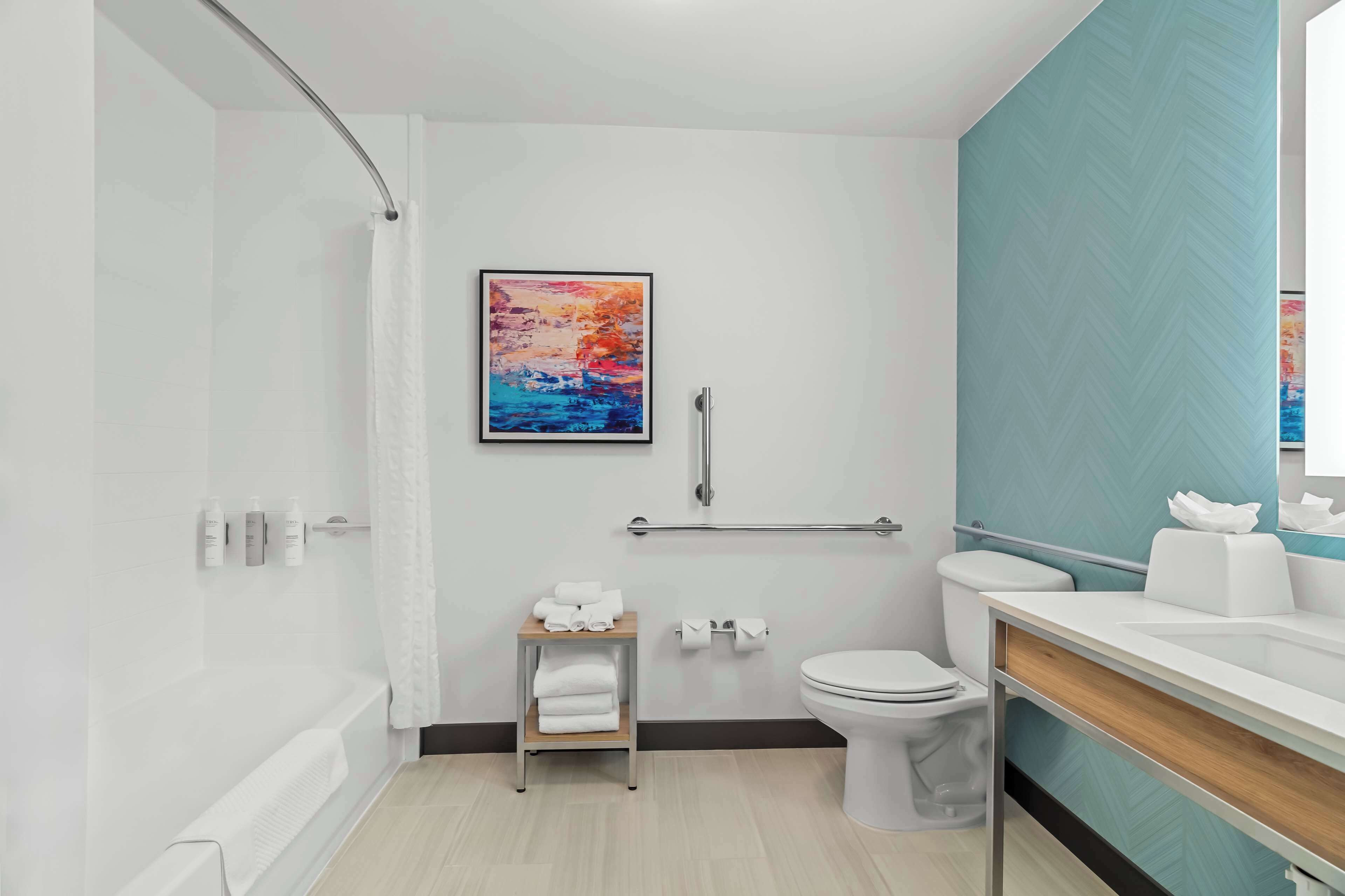 A modern bathroom with a shower, toilet, and sink. There is a towel rack and a framed painting on the wall.