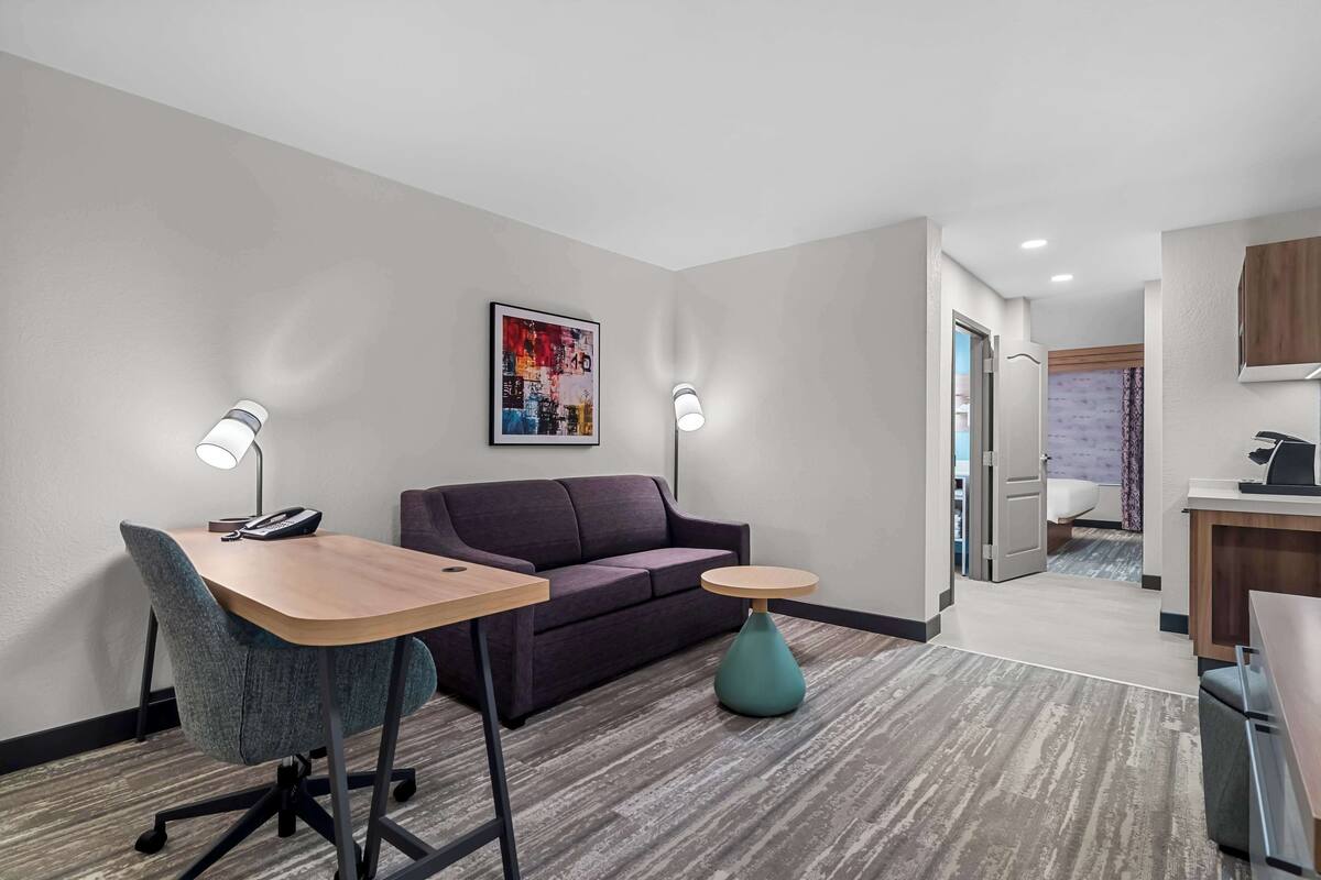 A hotel room with a desk, chair, sofa, and a small table with a lamp. There is a painting on the wall and a kitchen area visible through an open door.