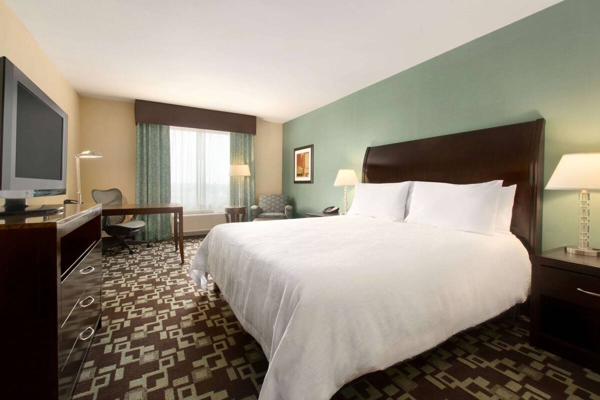 A hotel room with a large bed, a desk with a computer, a chair, a TV, and a window with curtains.