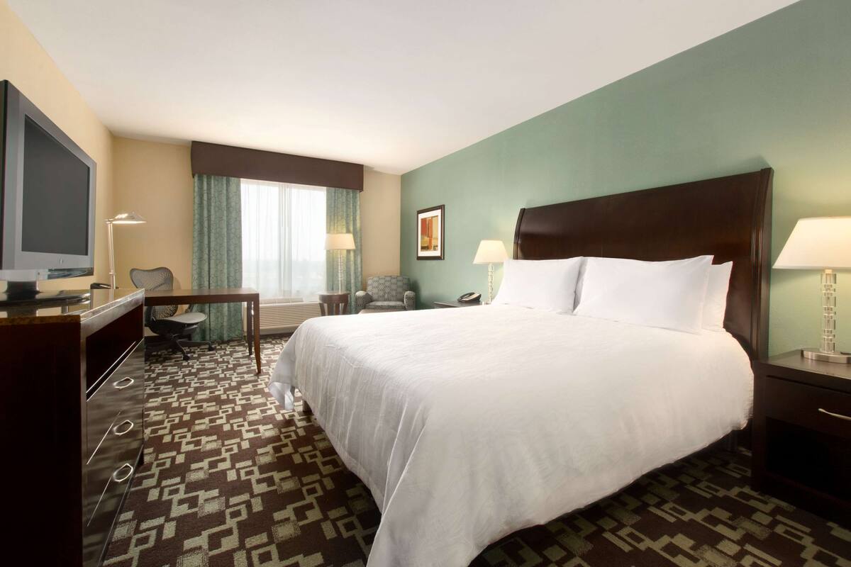 A hotel room with a large bed, a desk with a television, a chair, a lamp, and a window with curtains.