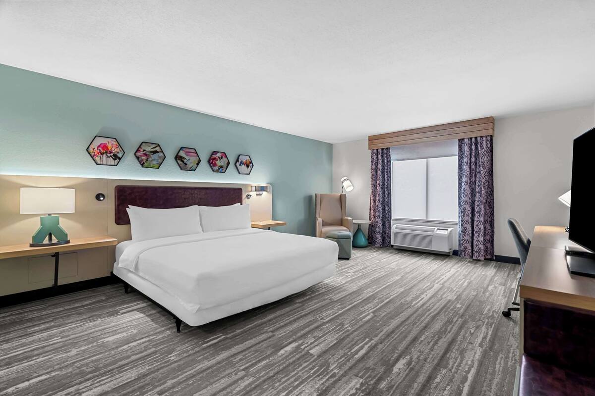A hotel room with a large bed, a desk with a computer, a chair, a window with curtains, and a wall-mounted TV.