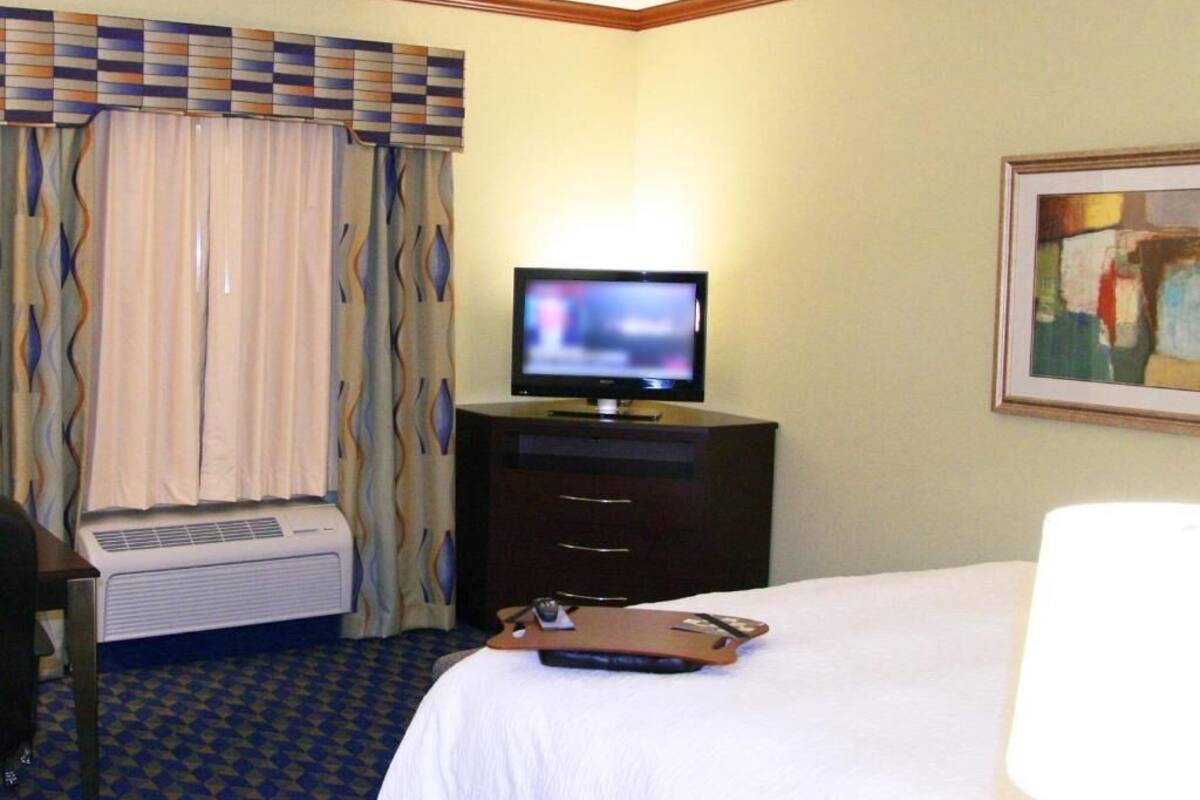 Ein Hotelzimmer mit Bett, Fernseher, Schreibtisch mit Stuhl, Schreibtischlampe und Wandklimaanlage.