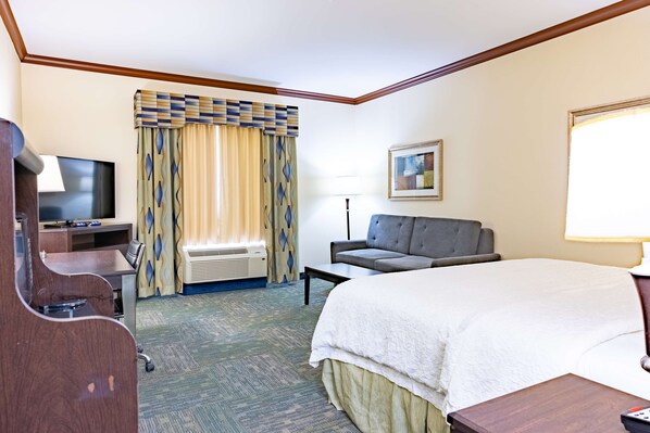 Quarto de hotel com uma cama, uma escrivaninha com computador, uma cadeira, uma televisão e uma janela com cortinas.
