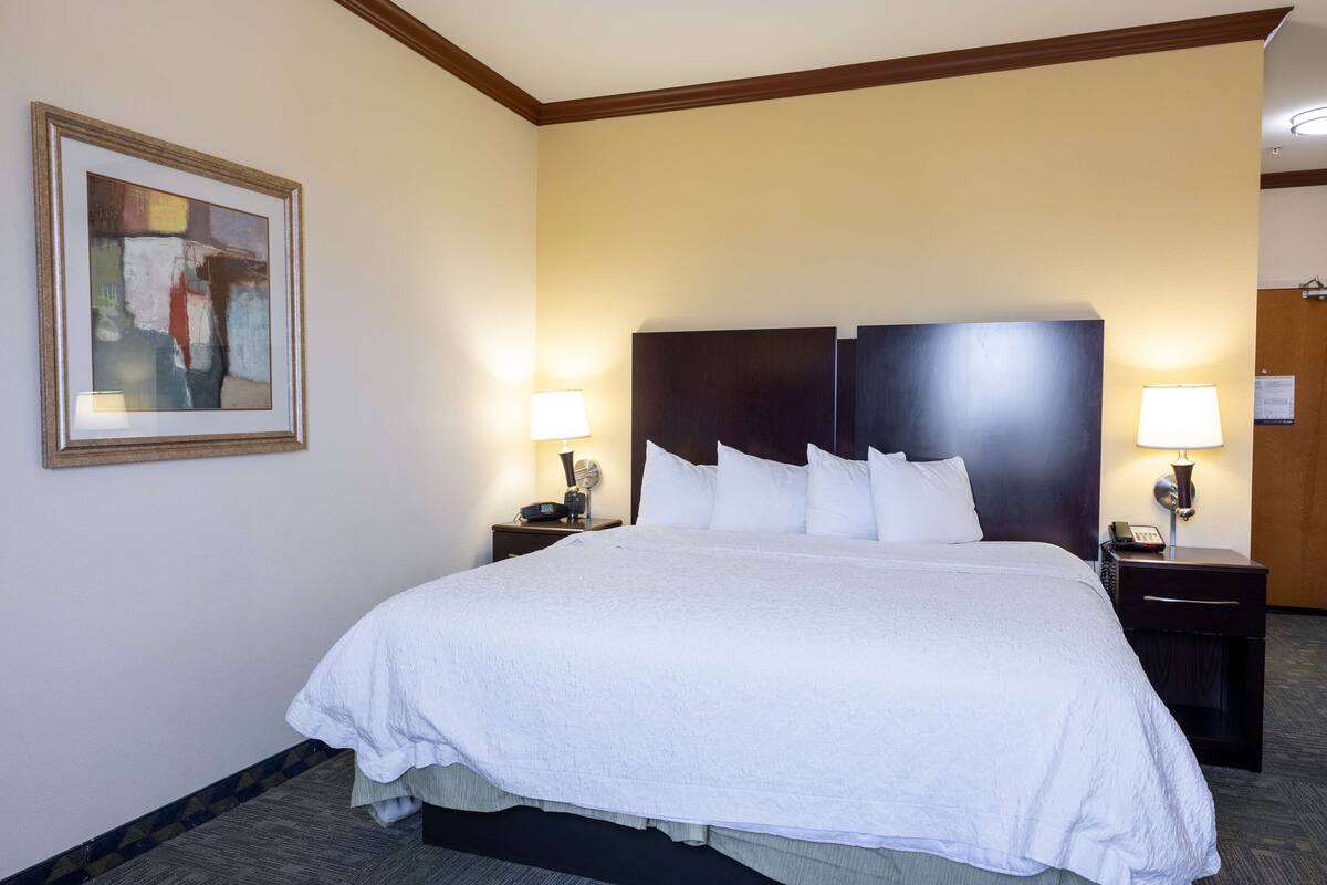 Une chambre d’hôtel avec un grand lit, deux tables de chevet avec des lampes, un tableau abstrait encadré au mur et une tête de lit en bois.