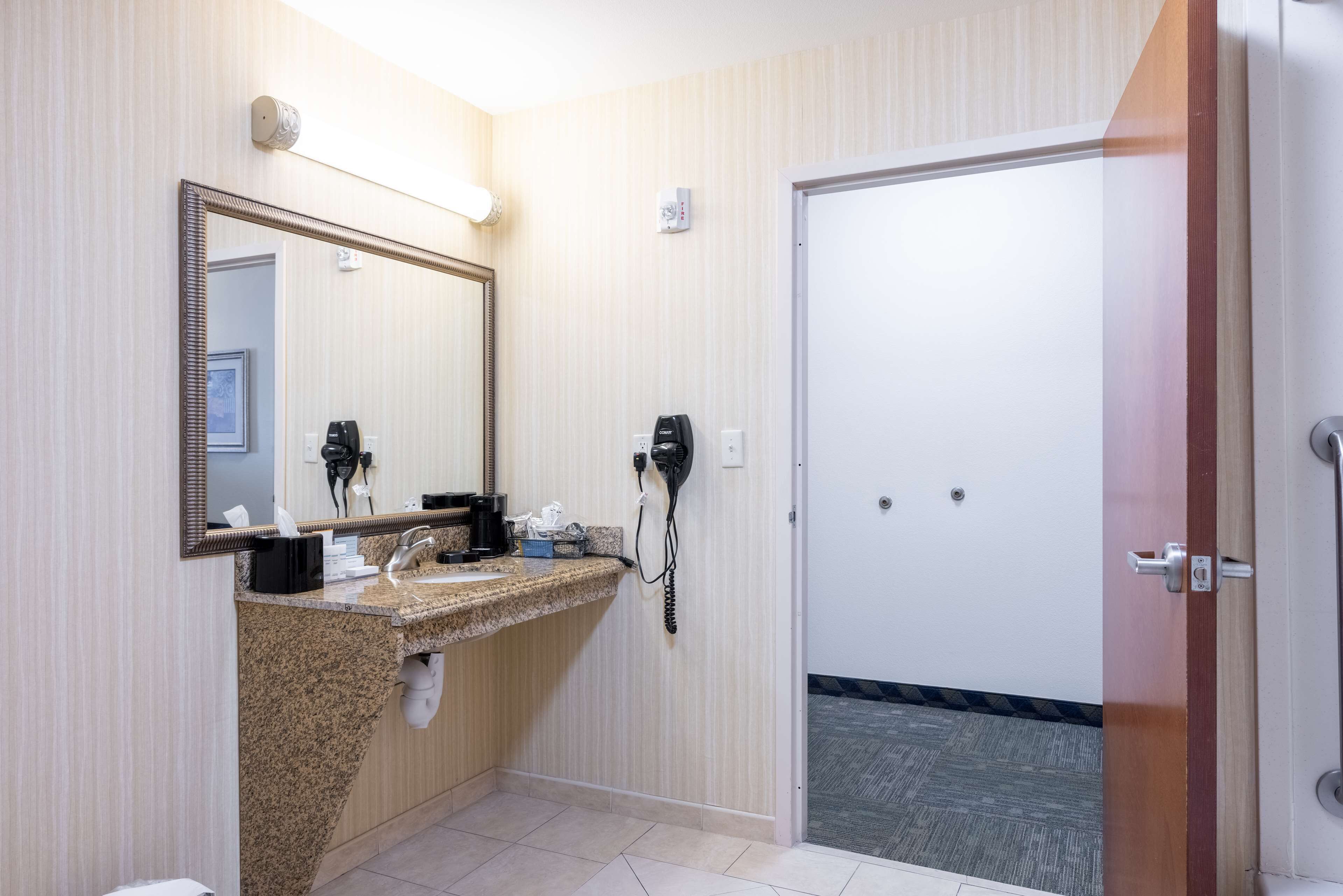 A hotel bathroom with a granite countertop, a large mirror, a hairdryer, and a door leading to a shower area.
