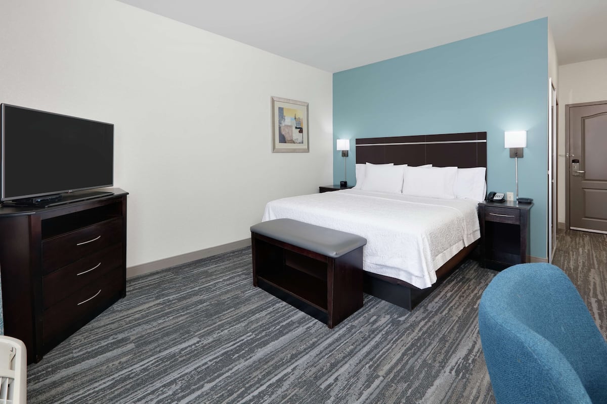 A hotel room with a bed, a desk with a flat-screen TV, a bench, a nightstand, and a chair.