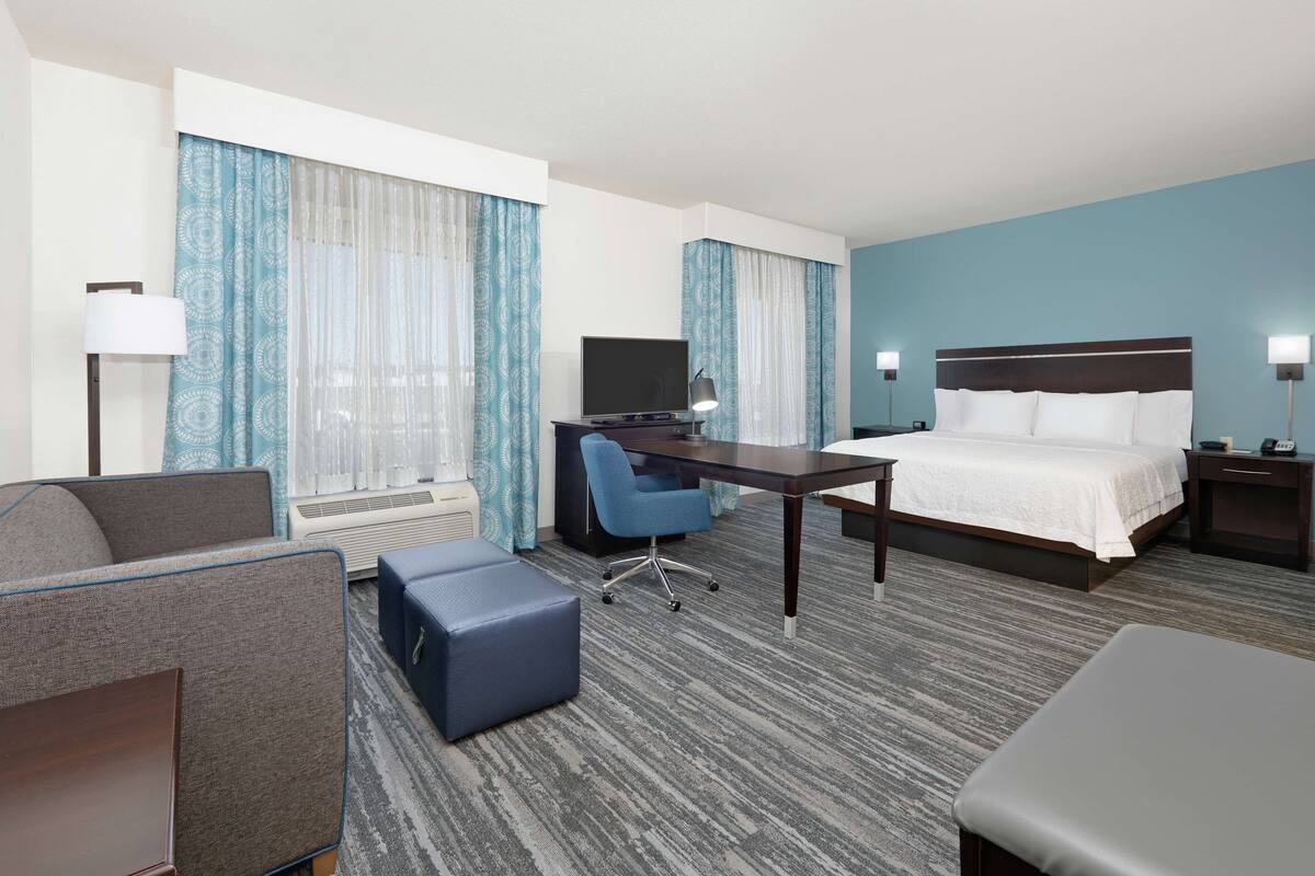 A hotel room with a bed, desk, chair, and a large window with curtains.