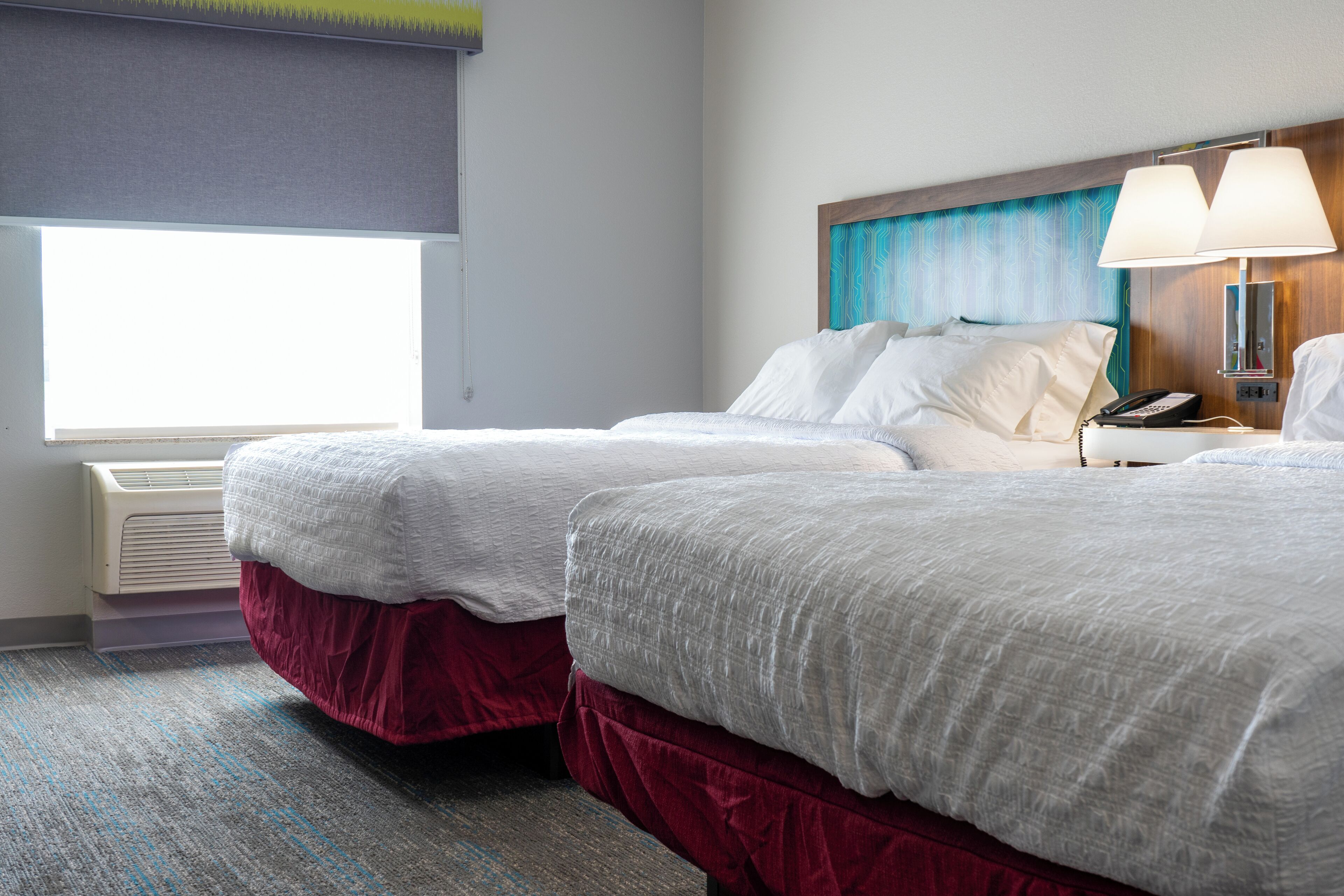 A hotel room with two beds, a window with blinds, a wall-mounted lamp, and a phone on the nightstand.