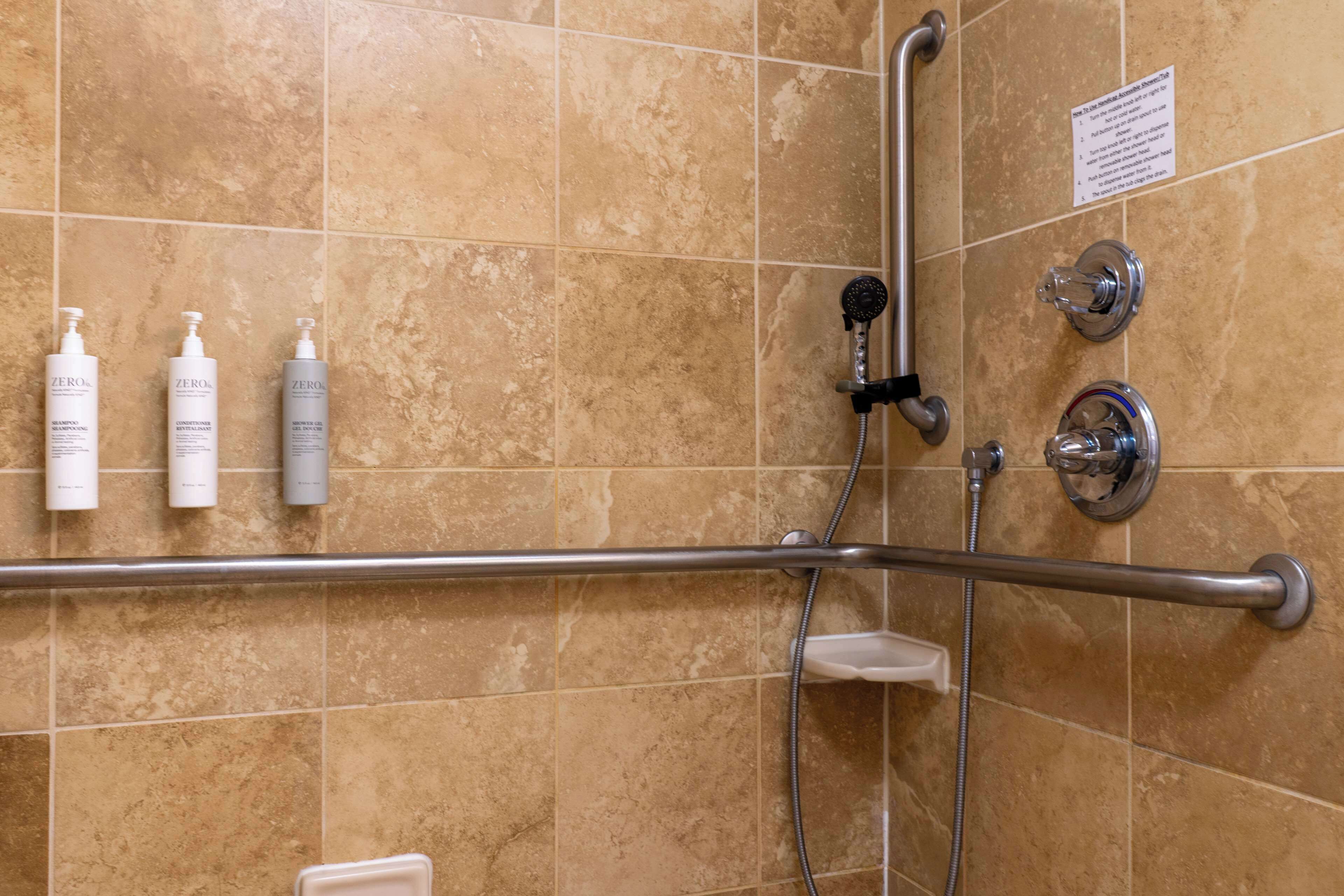 Baño con azulejos color crema, barra de apoyo, ducha con cabezal de mano y tres botellas de champú.