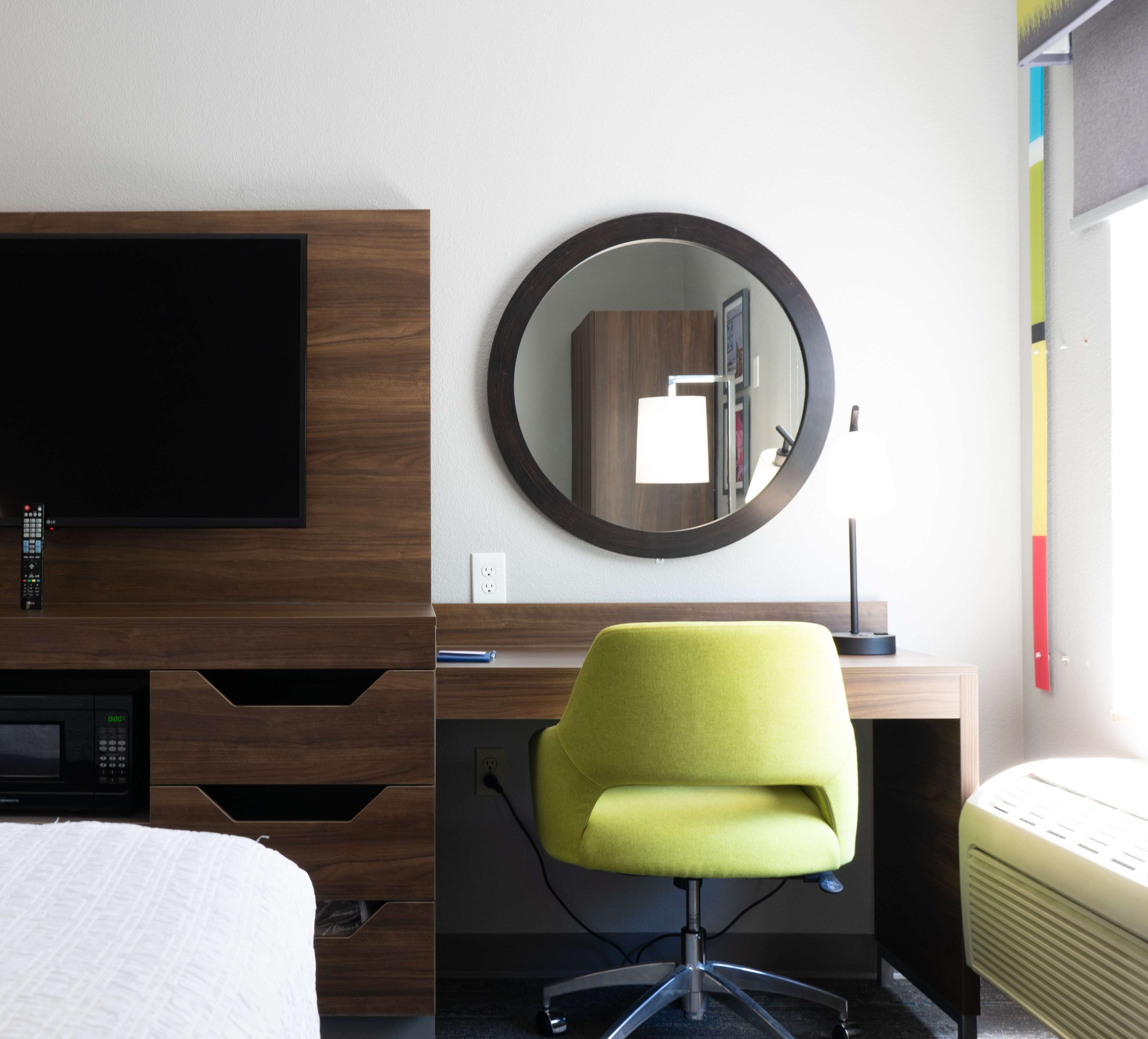A hotel room with a bed, a desk with a chair, a TV, a mirror, and a wall socket.