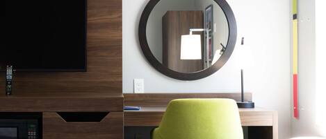 A hotel room with a bed, a desk with a chair, a TV, a mirror, and a wall socket.