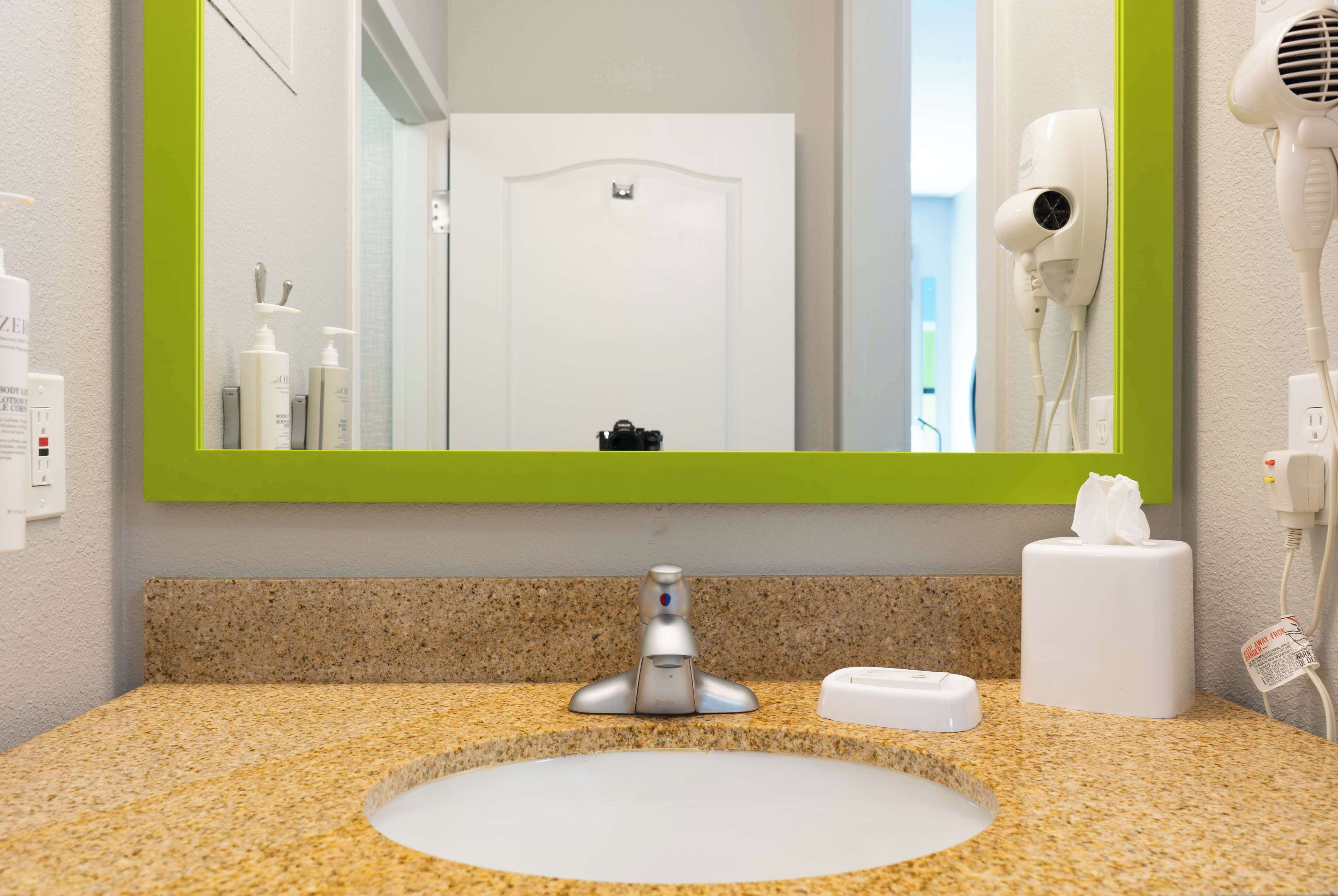 A bathroom with a granite countertop, a white sink, a green-framed mirror, a hairdryer, and a tissue dispenser.