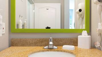 A bathroom with a granite countertop, a white sink, a green-framed mirror, a hairdryer, and a tissue dispenser.