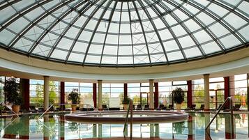 Ein großes Hallenbad mit Glasdach, umgeben von Liegestühlen und Topfpflanzen.