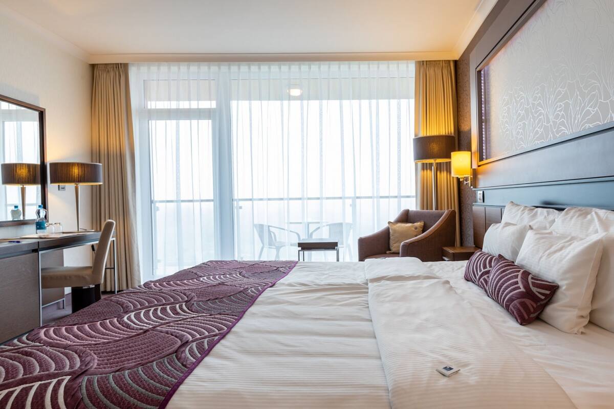A hotel room with a large bed, a desk with a chair, a small table, and a chair. There is a window with curtains and a view of the outside.