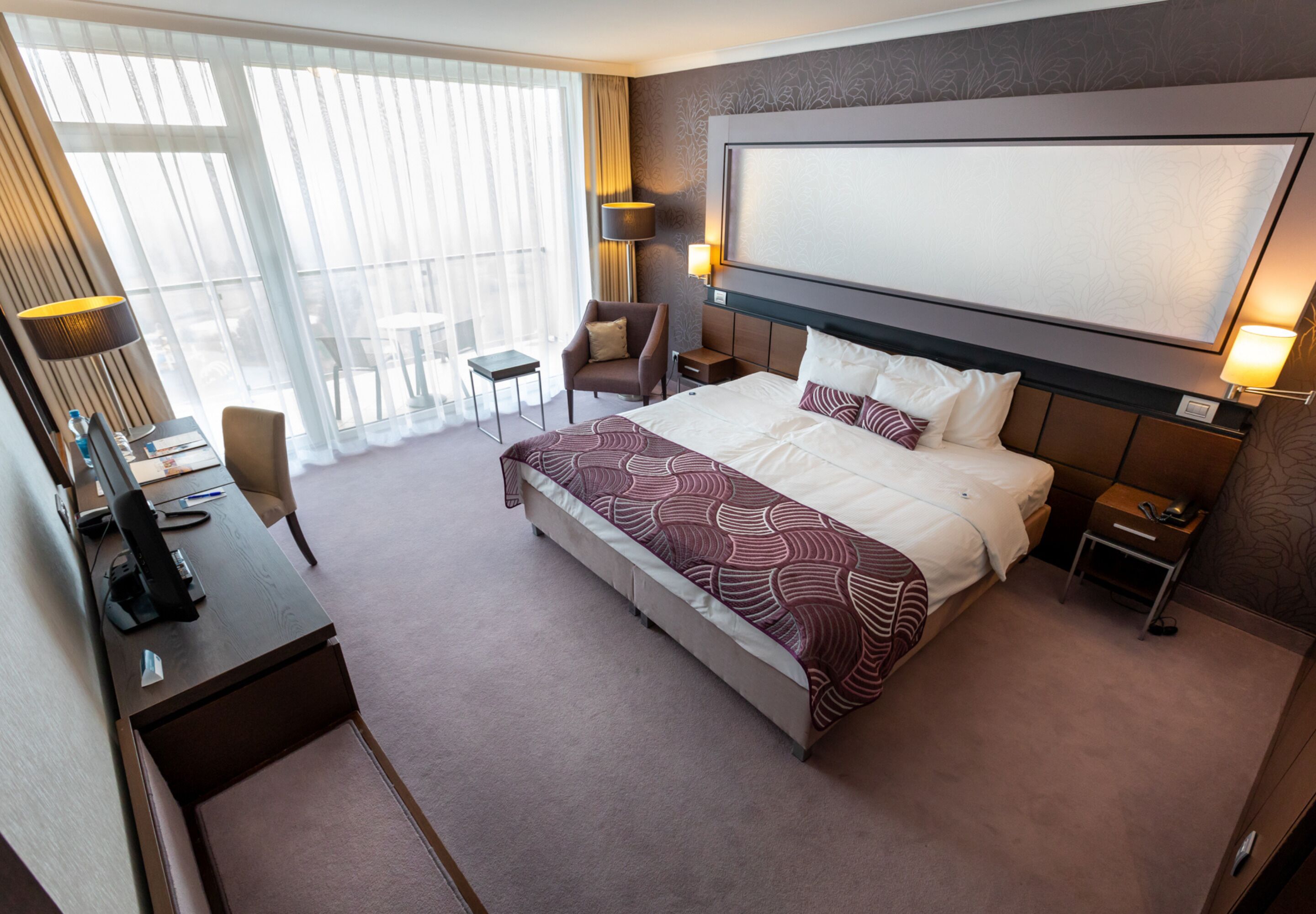 Una habitación de hotel con una cama grande, un escritorio con computadora, una silla, una mesita y una ventana con cortinas.