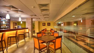 Un vestíbulo de hotel con zona de bar, varias mesas y sillas, y un separador de vidrio.