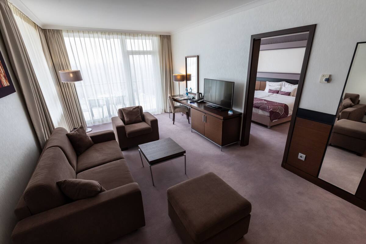 A hotel room with a sofa, armchair, ottoman, desk with a TV, and a bed.