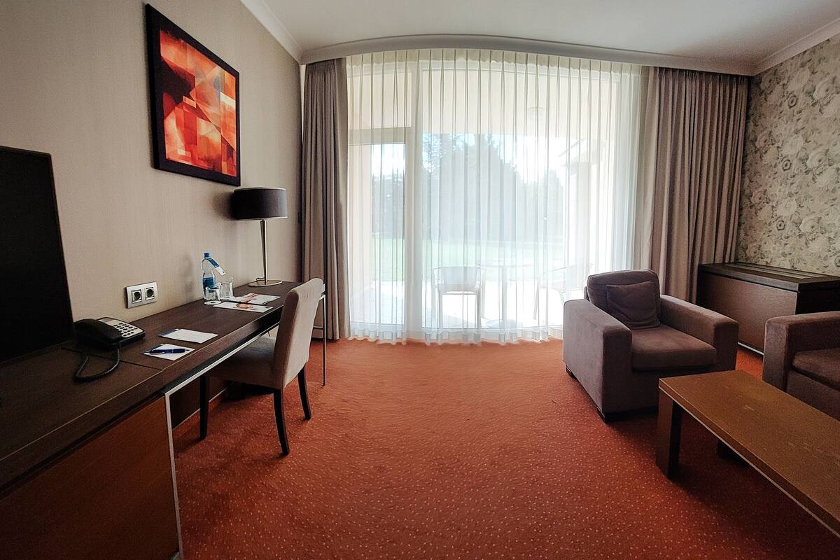 A hotel room with a desk, a chair, a sofa, and a large window with sheer curtains.