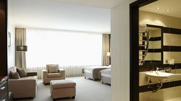 A hotel room with a sofa, armchair, ottoman, and a bed. There is a bathroom with a sink and mirror.