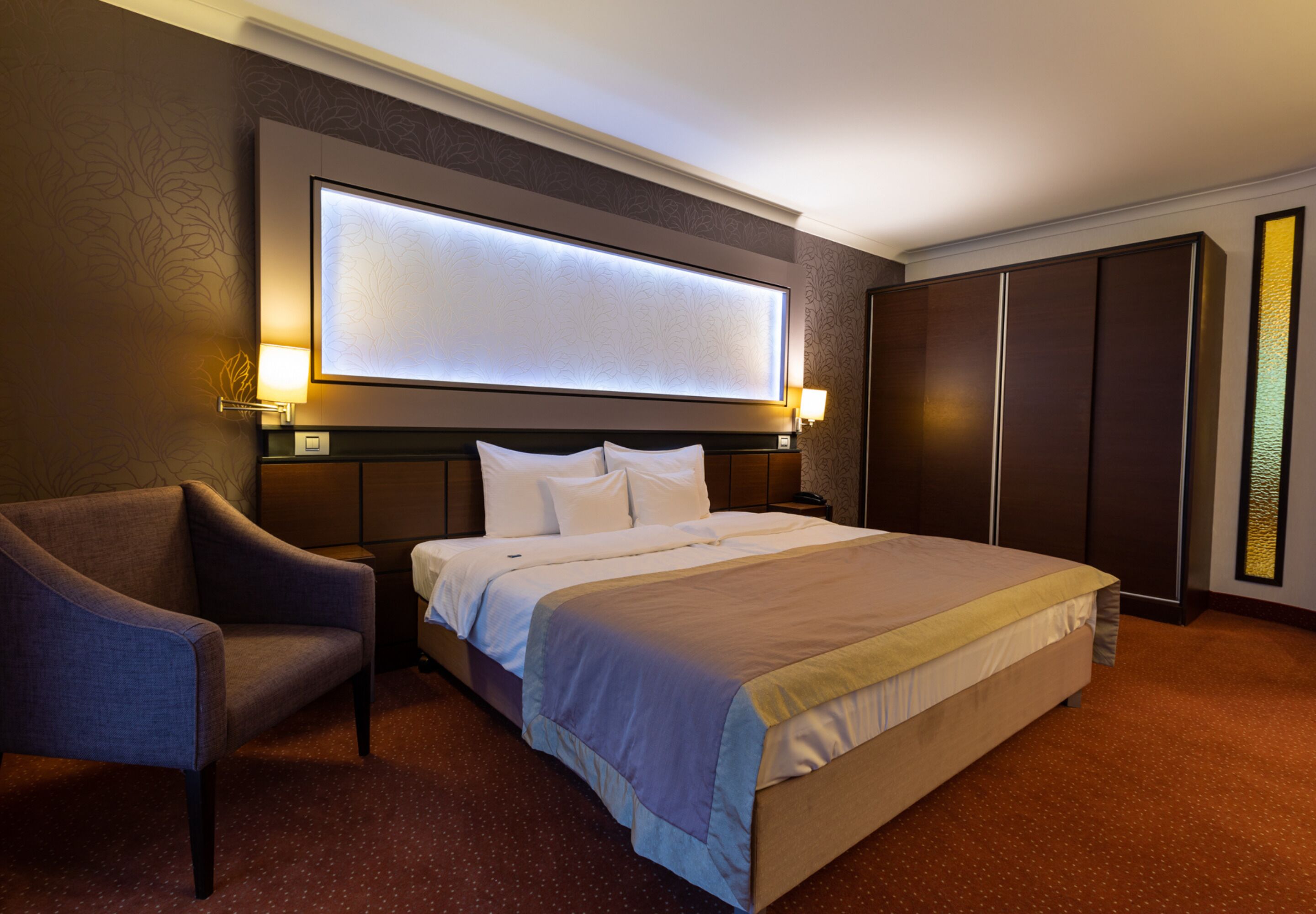 A hotel room with a large bed, a chair, a wardrobe, and a wall-mounted light fixture.