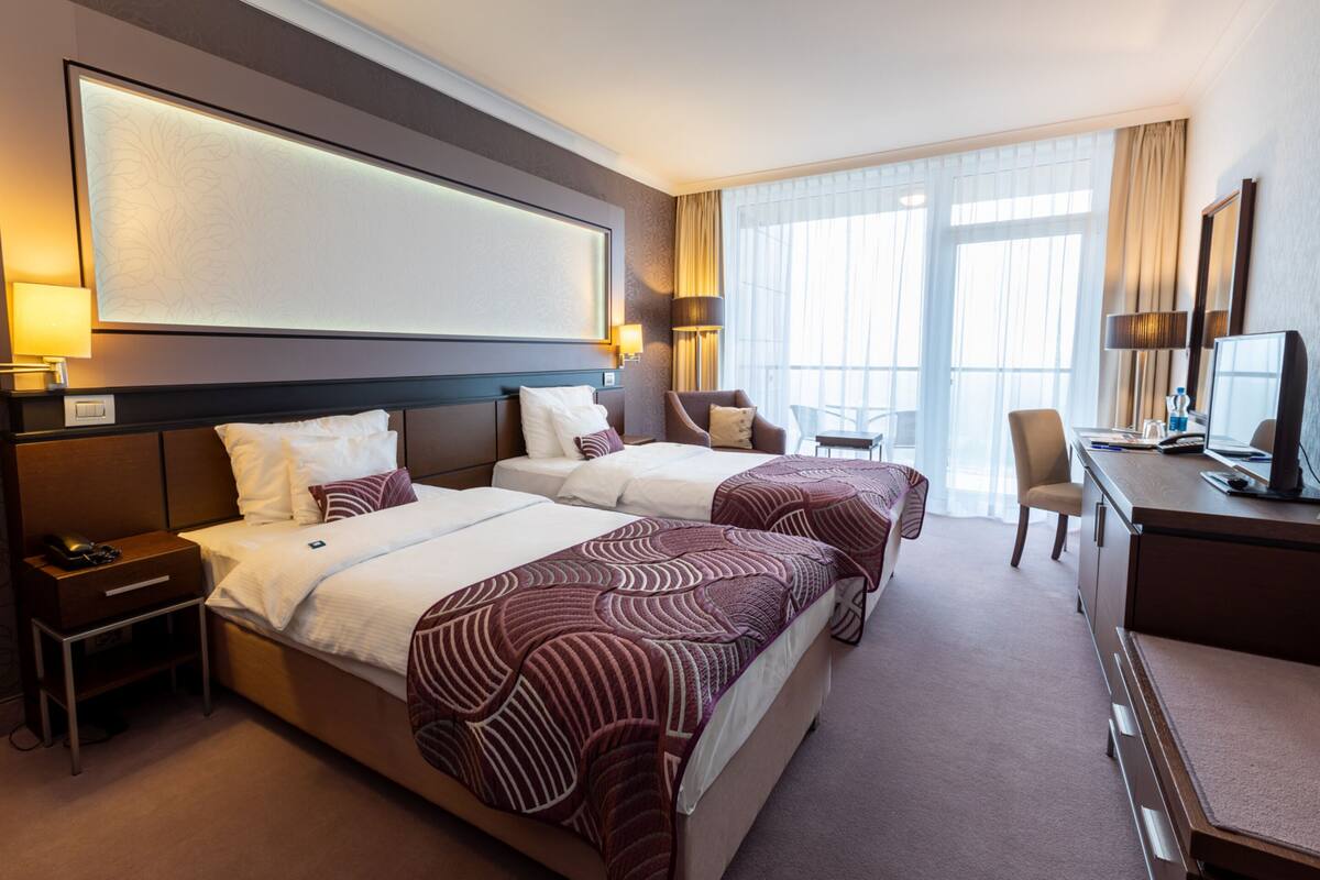 A hotel room with two beds, a desk, a chair, and a large window with curtains.