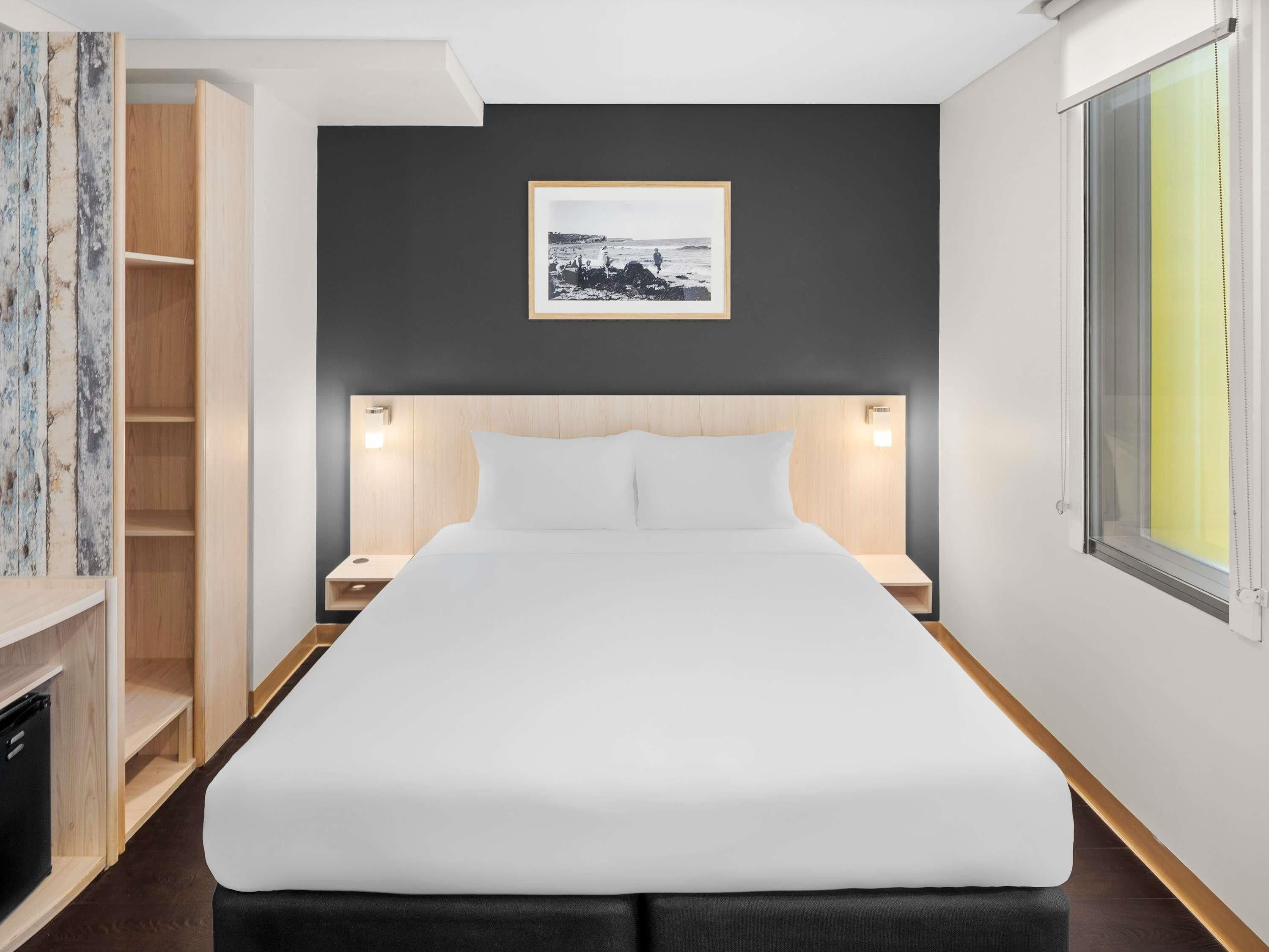 A modern hotel room with a large bed, wooden headboard, bedside tables, a framed picture on the wall, and a window with a view.