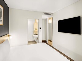Habitación de hotel con cama, televisor montado en la pared, baño con espejo y lavamanos, y un cuadro enmarcado en la pared.