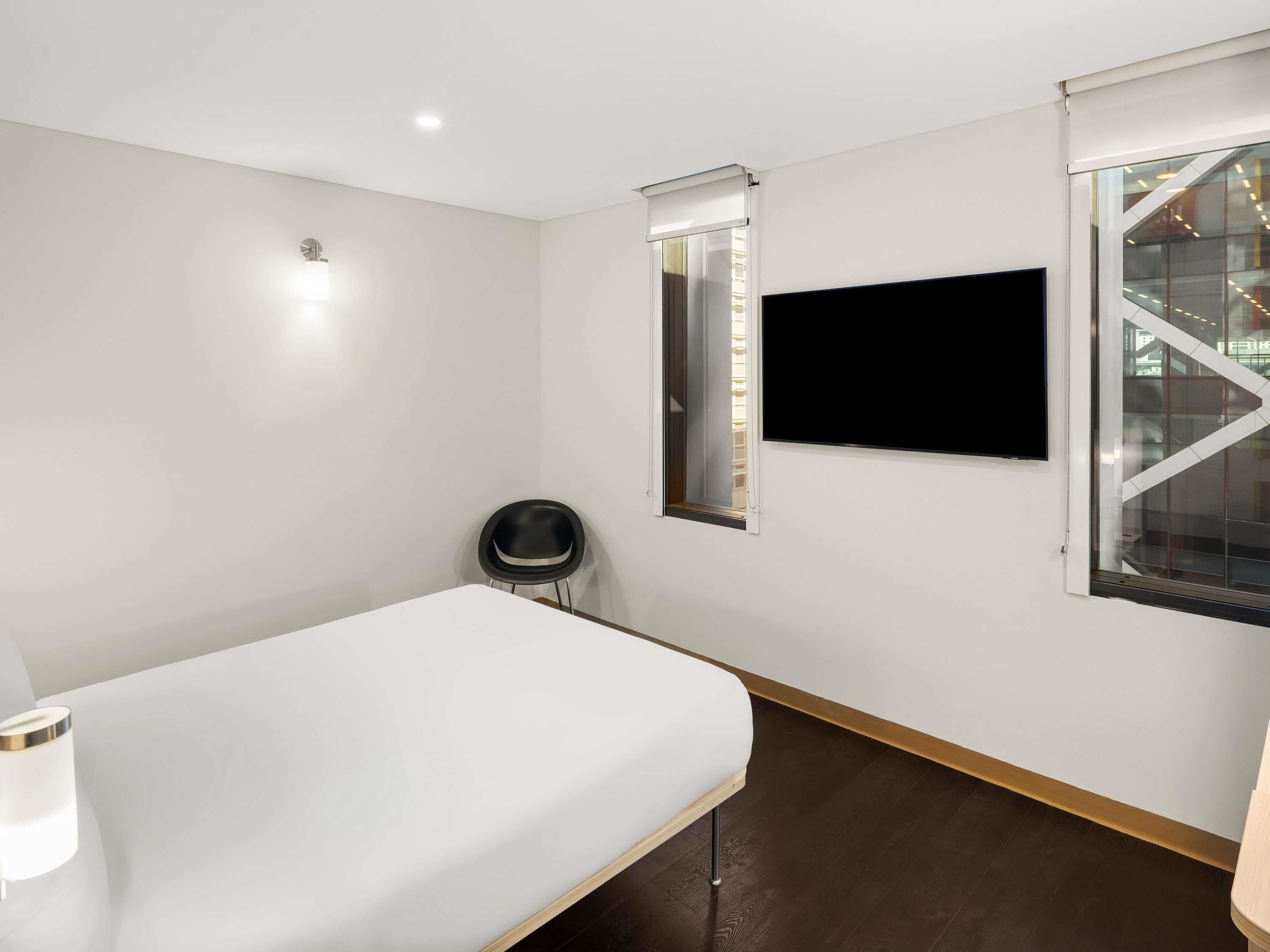 Une chambre d’hôtel moderne équipée d’un lit, d’une chaise, d’une télévision et d’une fenêtre donnant sur un immeuble.