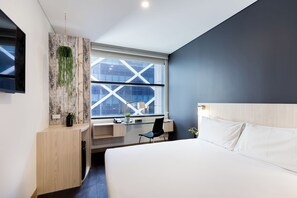 A modern hotel room with a large bed, a desk with a chair, a window with a view of the city, and a wall-mounted TV.