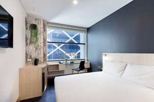 Quarto de hotel moderno com uma cama grande, uma escrivaninha com cadeira, uma janela com vista para a cidade e uma TV fixada na parede.