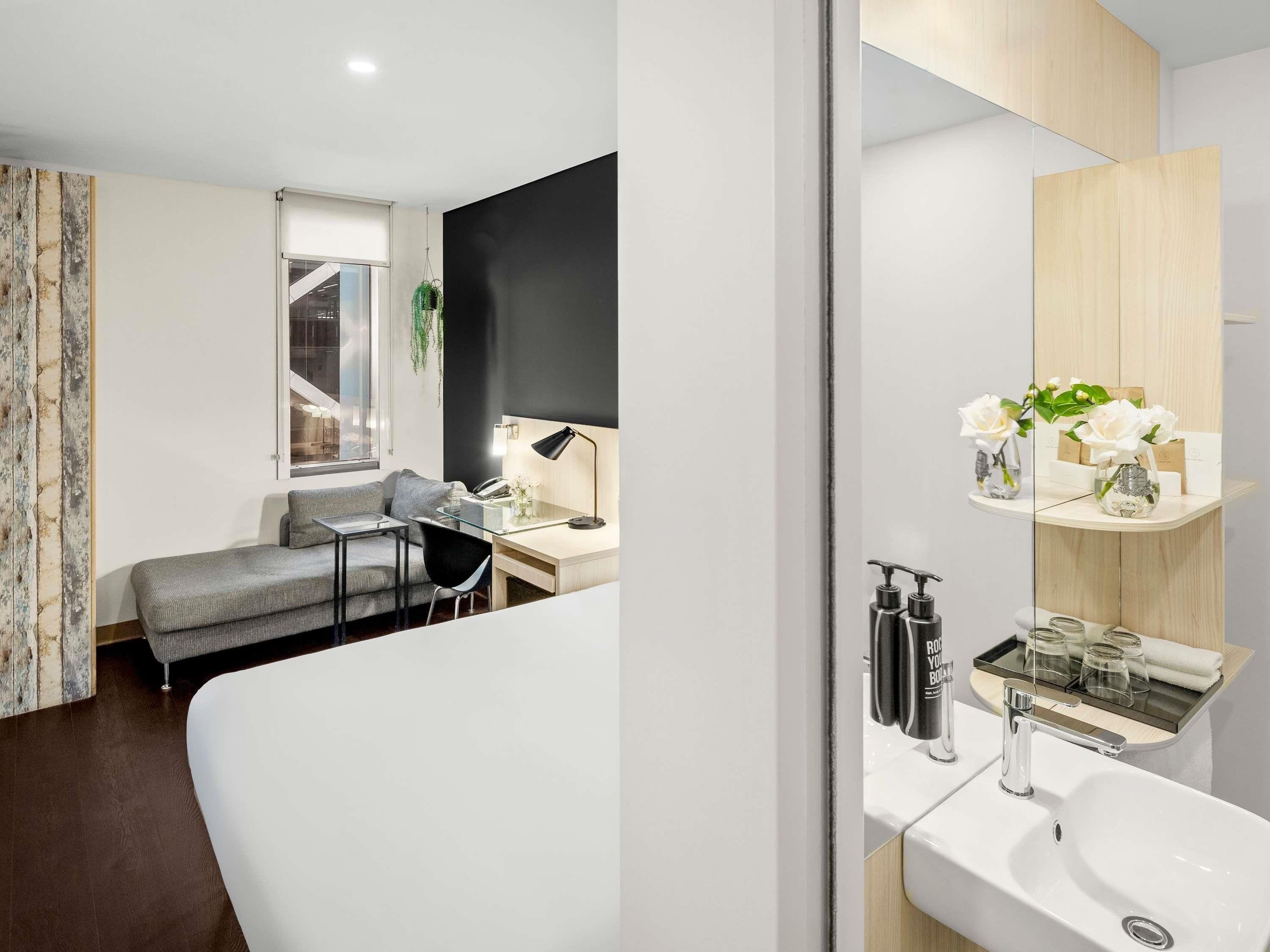 A modern hotel room with a curved bed, a desk with a lamp, a small seating area, and a bathroom with a mirror, sink, and decorative items.