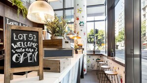 A modern bar area with a chalkboard sign reading "LOT 51 WELCOME reles are OPEN", wooden crates, hanging plants, and a view of the street through large windows.