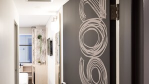 Un pasillo moderno de hotel con una puerta oscura que presenta un diseño abstracto blanco, que conduce a un baño con ducha y lavamanos.