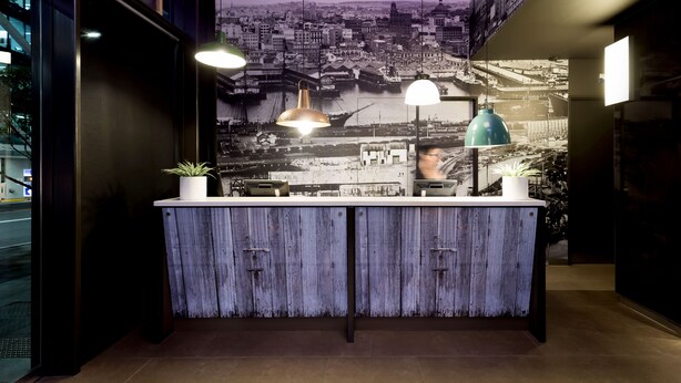 A reception desk with a cityscape mural, pendant lighting, and a potted plant. - ibis Sydney Barangaroo Hotel (Sydney)