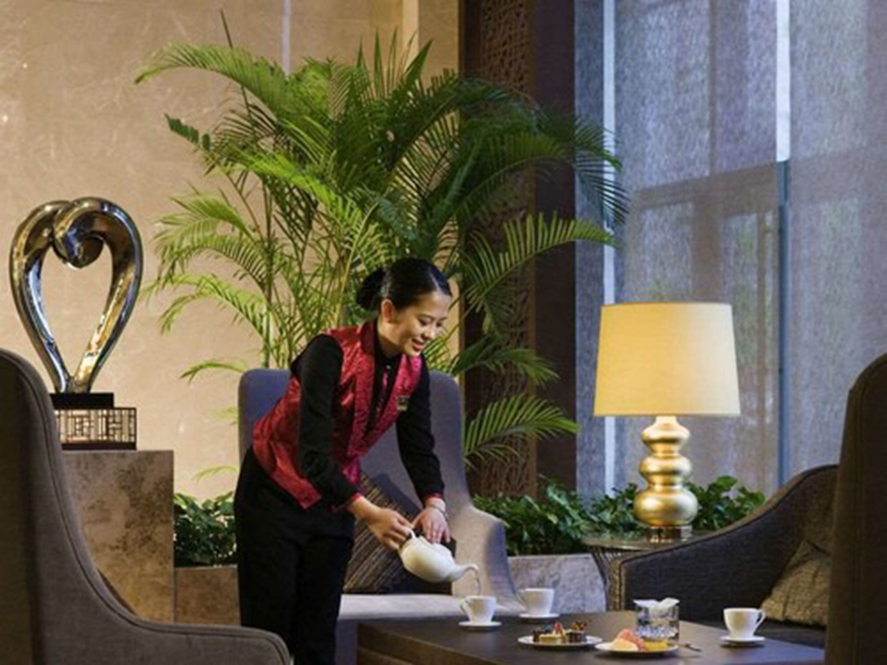 A hotel lobby with a reception desk, a large potted plant, a heart-shaped sculpture, and a table with a lamp and teapot.