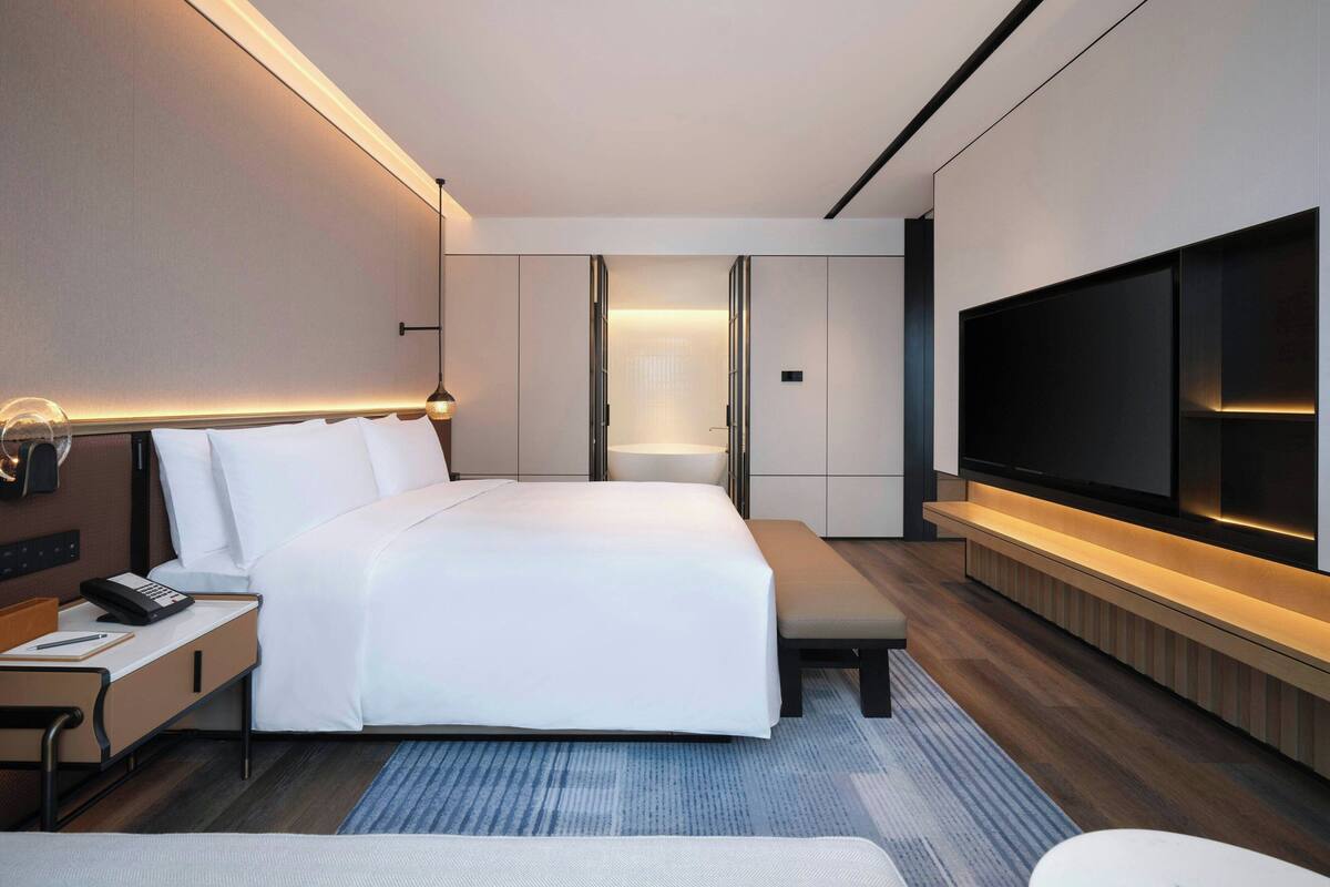 Quarto de hotel moderno com uma cama grande, mesa de cabeceira com telefone, TV de tela plana na parede e um banco.