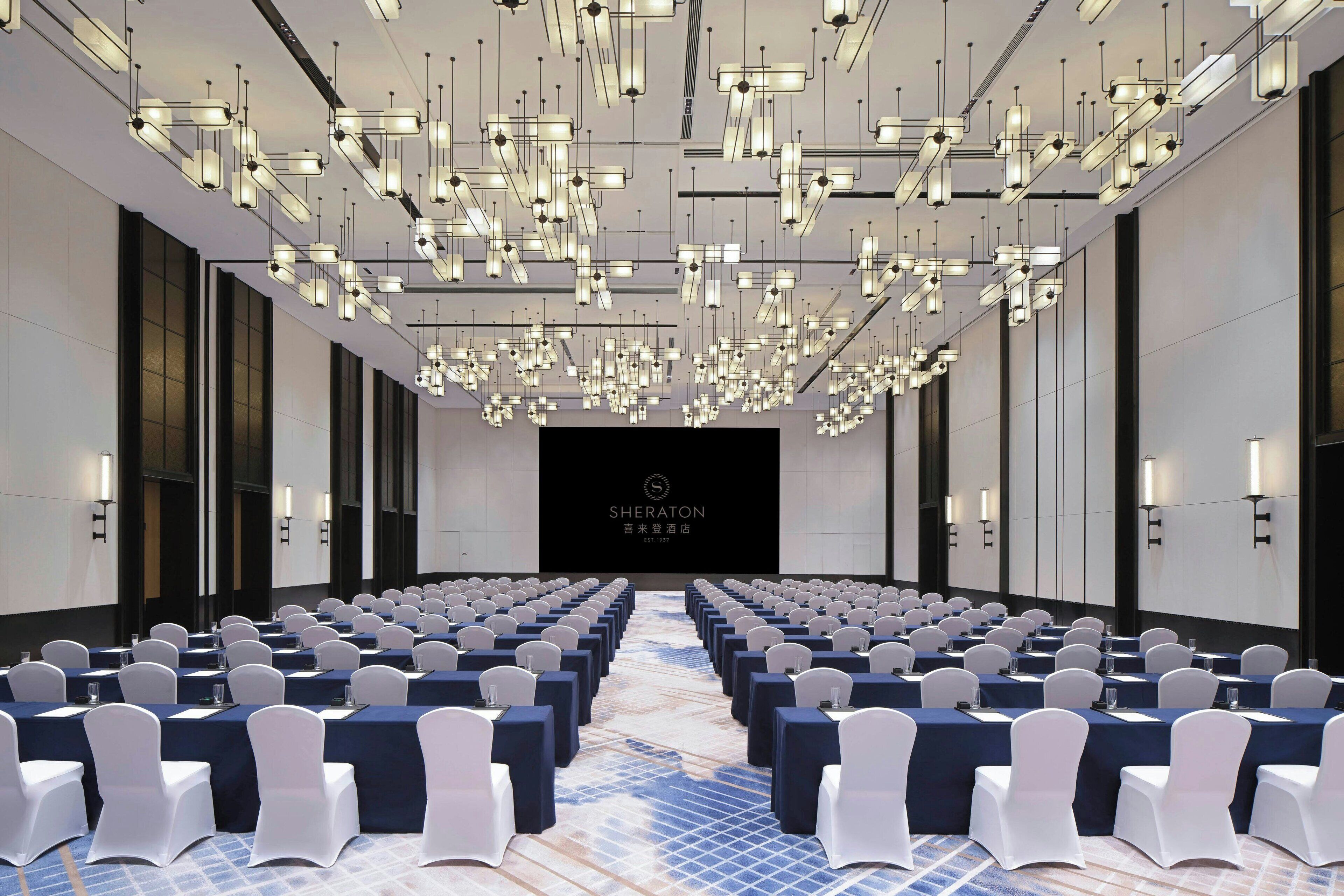 A conference room with rows of white chairs, a stage with a screen, and modern lighting fixtures.