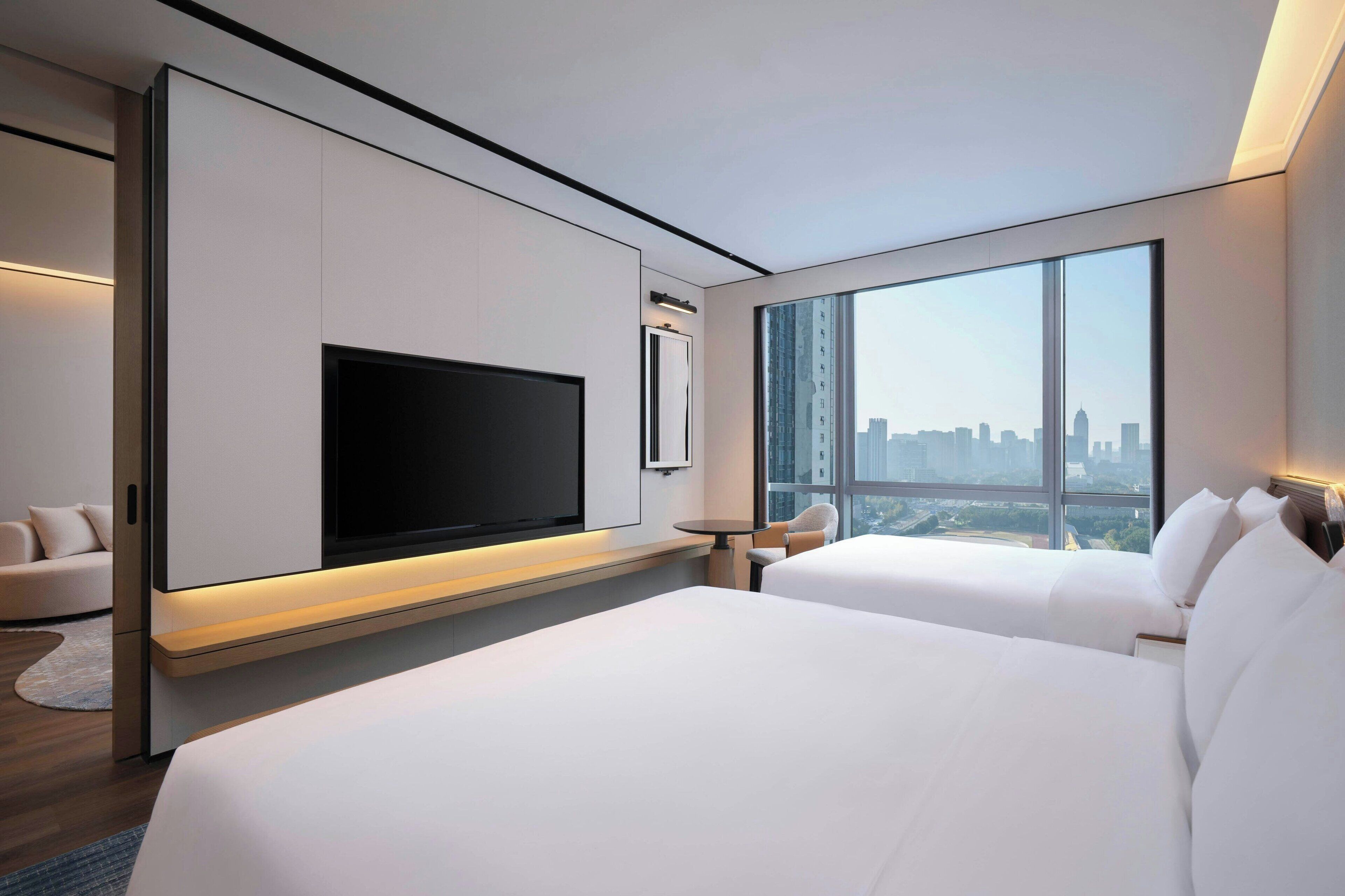 A modern hotel room with a large bed, a flat-screen TV, a city view, and a minimalist design.