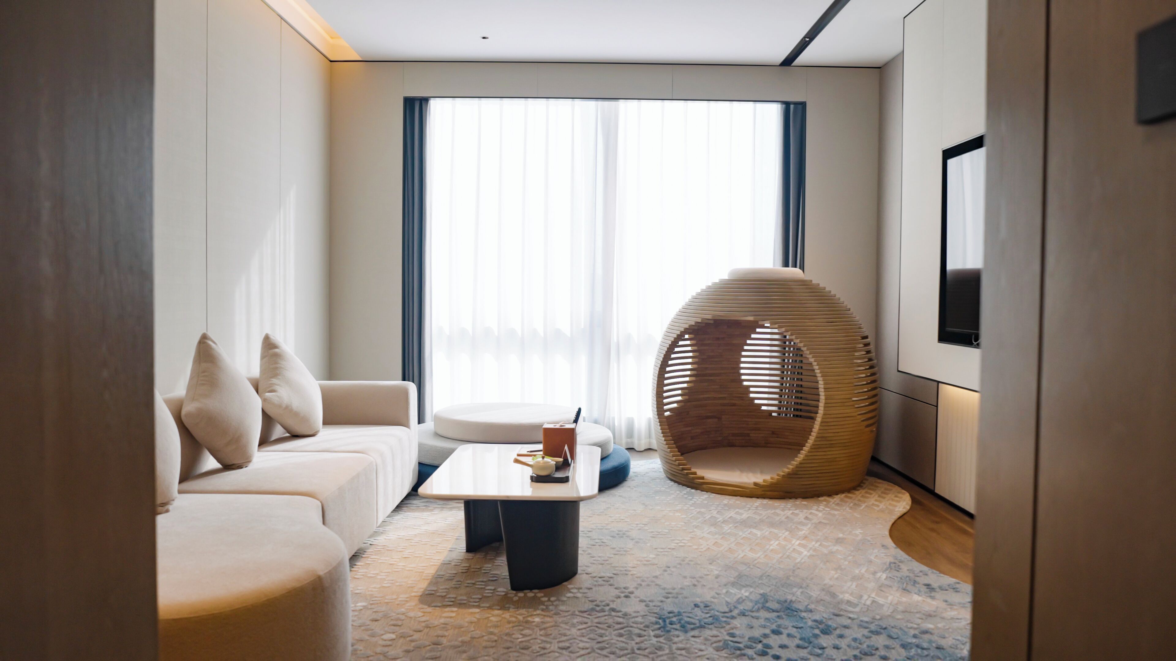 A modern hotel room with a sofa, a coffee table, a unique hanging chair, and a large window with sheer curtains.