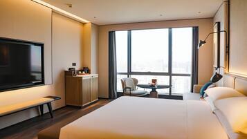 A hotel room with a large bed, a TV, a small table with a chair, a coffee station, and a view of the city through large windows.