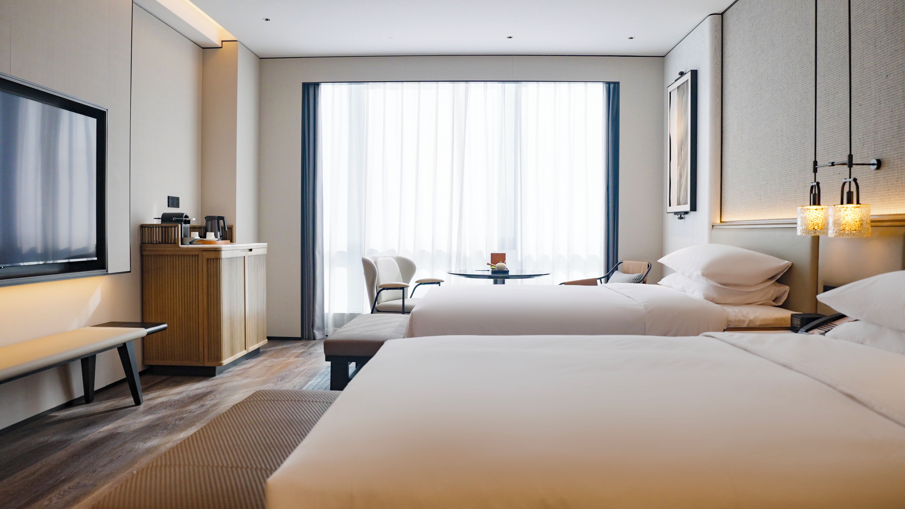 A hotel room with two beds, a desk, a chair, a coffee station, and a large window with curtains.