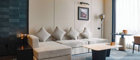 A modern hotel room with a sectional sofa, a small table, a floor lamp, and a large window.