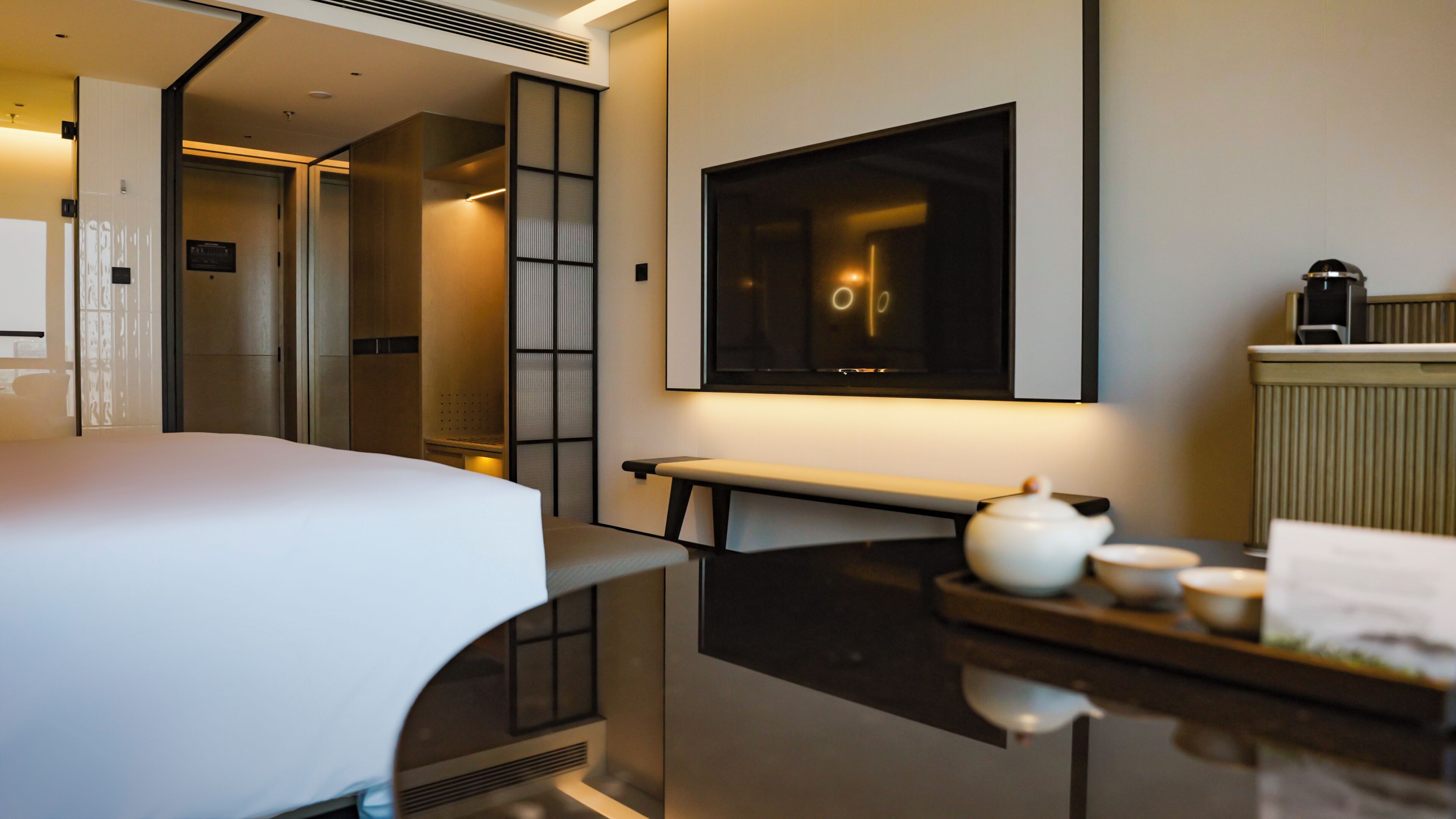 A modern hotel room with a bed, a small desk with a teapot, a mirror, and a view of a corridor.