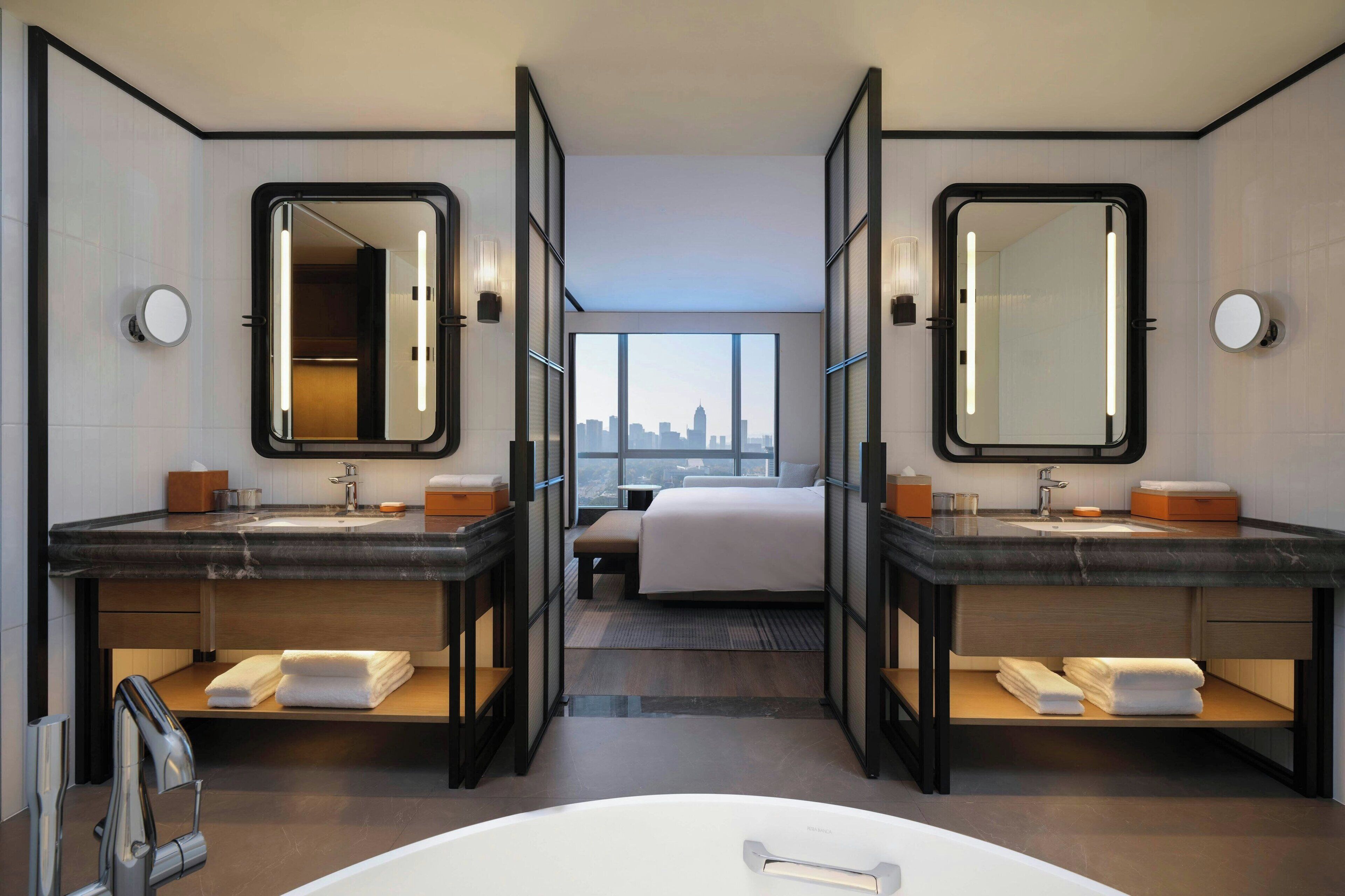 A modern hotel room with two bathrooms, a large bed, and a city view.