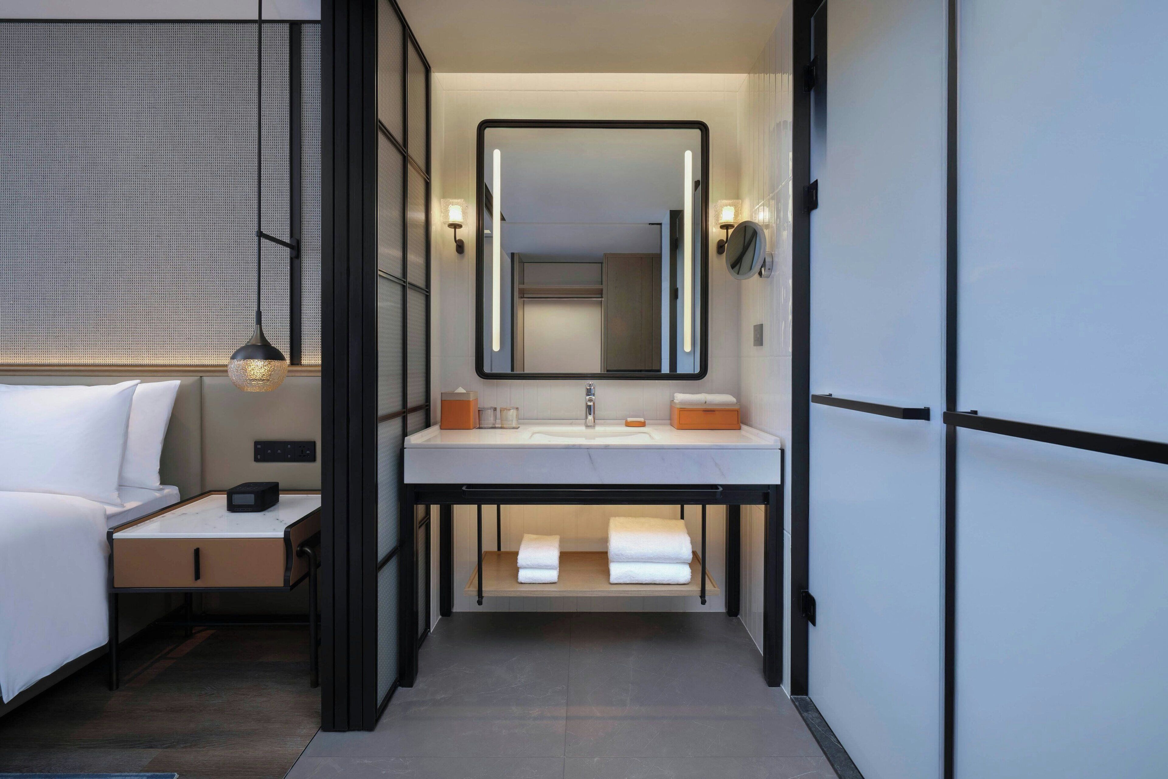 A modern bathroom with a large mirror, a sink, and a bedside table with a lamp.