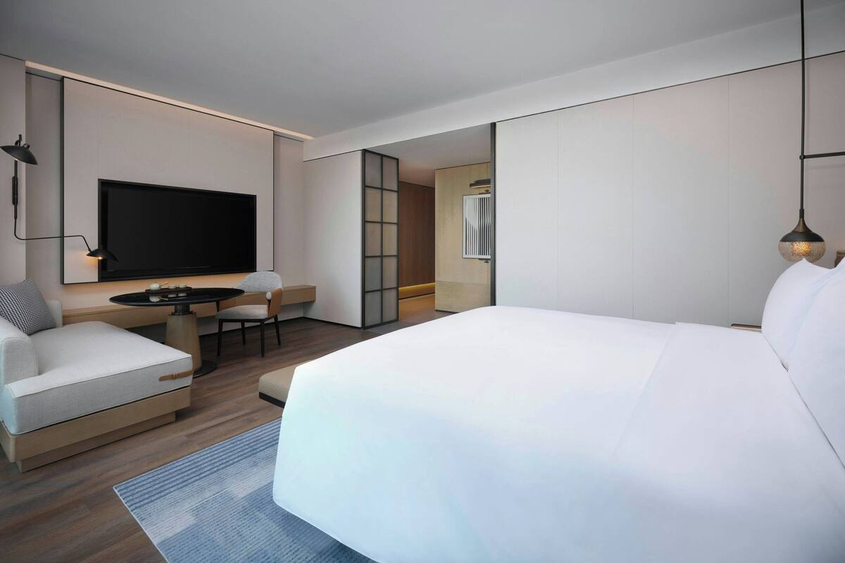 Quarto de hotel moderno com uma cama grande, televisão de tela plana, escrivaninha com cadeira e um pequeno espaço para sentar.