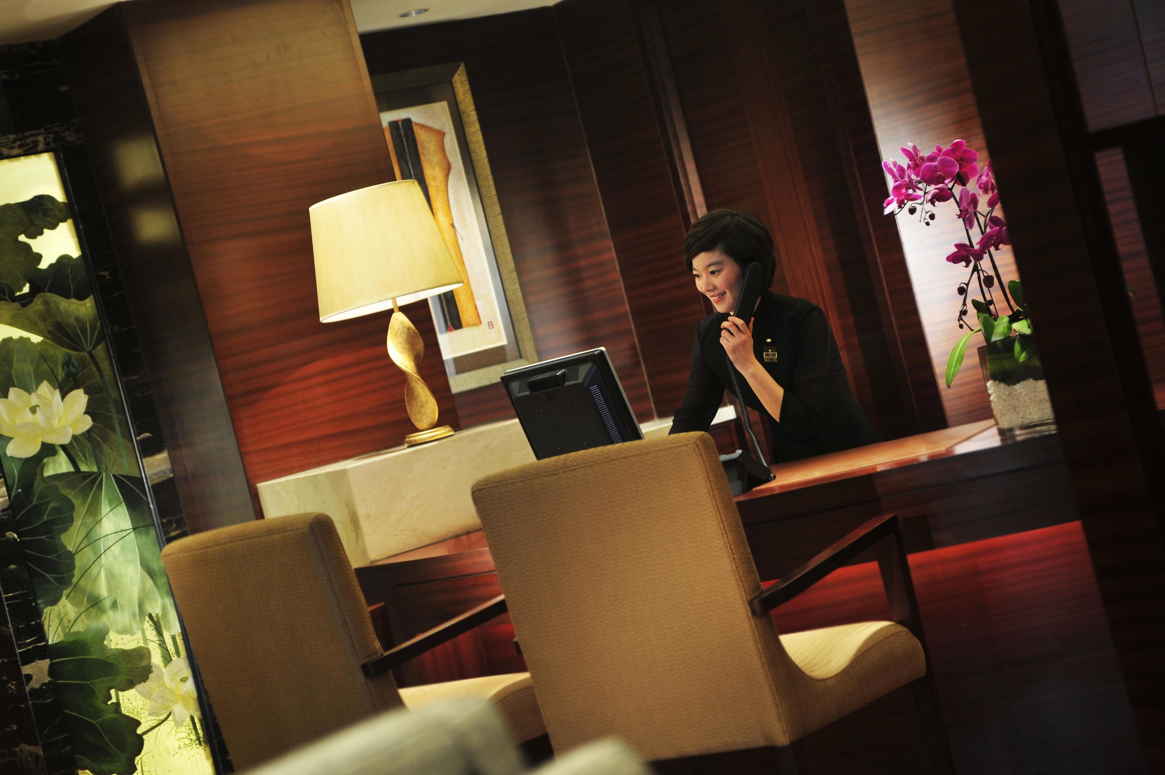 Un'area reception di un hotel con una recezionista, una scrivania, sedie, una lampada e un'orchidea in vaso.