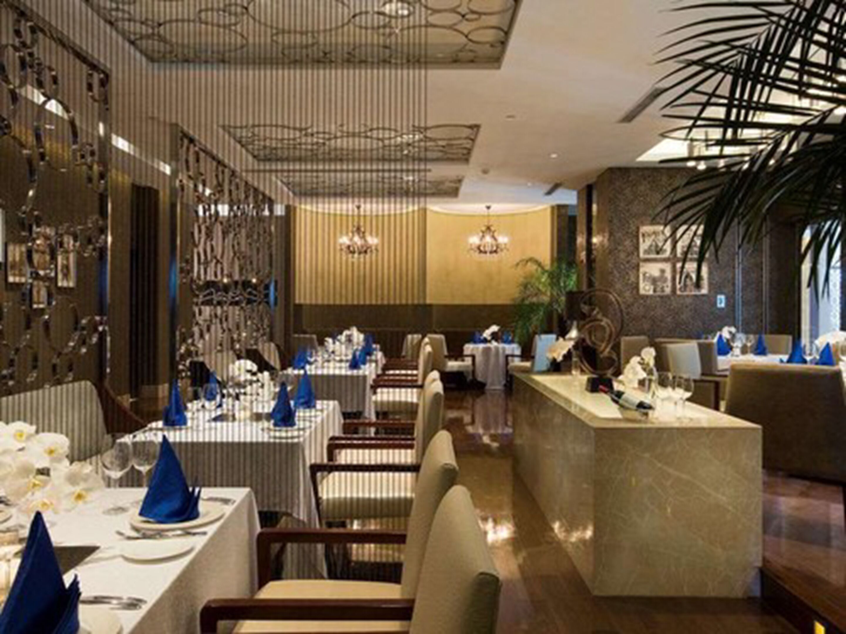A restaurant with multiple dining tables set with blue napkins and white tablecloths, a central bar area, and a chandelier.